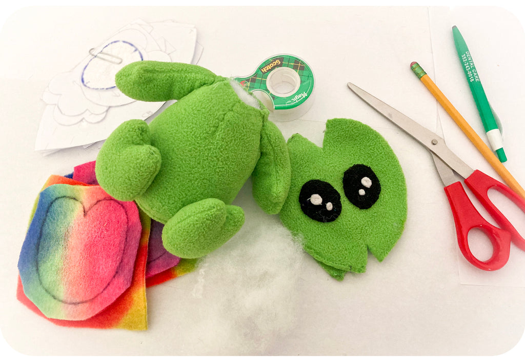 Alien plush laying on a craft table with fabric, stuffing, and scissors.